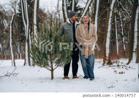 Couple enjoying winter outdoors, standing beside a small evergreen tree in a snowy forest, capturing the joy of the holiday season and nature's beauty 140014757