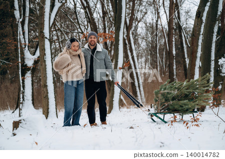 Couple enjoying winter outdoors while pulling a Christmas tree on a sled through a snowy forest, capturing the essence of holiday traditions and togetherness Couple enjoying winter outdoors while pulling a Christmas tree on a sled through a snowy forest, capturing the essence of holiday traditions and togetherness 140014782