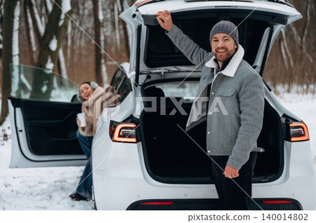 Smiling man in winter attire stands by open trunk of white car in snowy forest, while woman enjoys warm drink, showcasing joyful outdoor adventure Smiling man in winter attire stands by open trunk of white car in snowy forest, while woman enjoys warm drink, showcasing joyful outdoor adventure 140014802