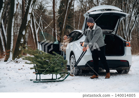 Man pulling a sled with a freshly cut Christmas tree while a woman sits in a car, surrounded by snow-covered trees in a winter landscape Man pulling a sled with a freshly cut Christmas tree while a woman sits in a car, surrounded by snow-covered trees in a winter landscape 140014805