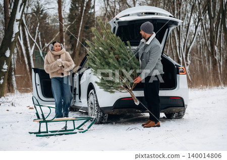 Couple enjoying winter day, unloading Christmas tree from car in snowy forest, with sled nearby, capturing festive holiday spirit and outdoor adventure 140014806