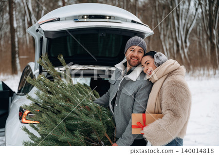 Couple joyfully loading a Christmas tree and gift into a car trunk, surrounded by a snowy winter landscape, capturing the festive holiday spirit and togetherness Couple joyfully loading a Christmas tree and gift into a car trunk, surrounded by a snowy winter landscape, capturing the festive holiday spirit and togetherness 140014818
