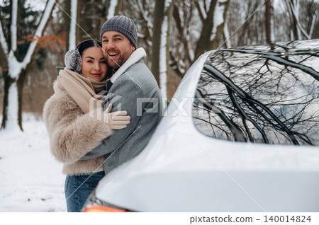 Couple embracing in winter attire, smiling joyfully beside a car in a snowy landscape, capturing the warmth of love and togetherness during the cold season 140014824