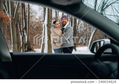 Couple embracing in winter forest, framed by car interior, showcasing love and warmth in a snowy landscape with trees in the background Couple embracing in winter forest, framed by car interior, showcasing love and warmth in a snowy landscape with trees in the background 140014828