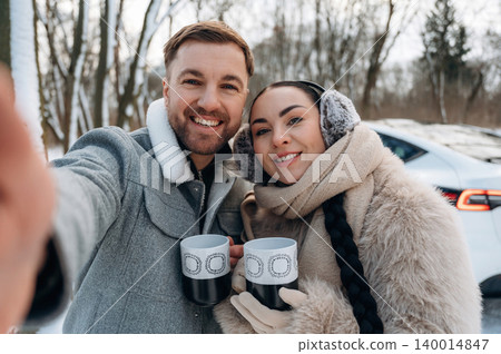 Smiling couple enjoying winter outdoors, holding mugs while taking a selfie, surrounded by snowy trees and a car in the background, capturing a joyful moment Smiling couple enjoying winter outdoors, holding mugs while taking a selfie, surrounded by snowy trees and a car in the background, capturing a joyful moment 140014847