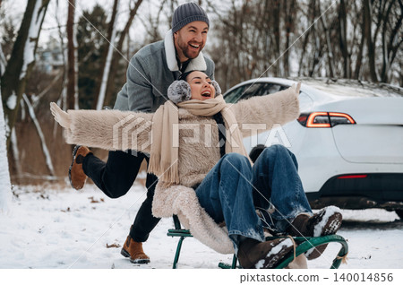 Joyful couple enjoying winter fun on sled in snowy landscape, with car parked nearby, capturing the essence of outdoor adventure and seasonal excitement 140014856