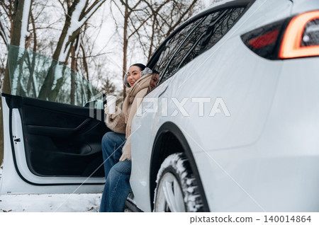 Woman enjoying warm beverage while sitting on snowy ground beside modern electric vehicle with open door, showcasing winter outdoor adventure and relaxation Woman enjoying warm beverage while sitting on snowy ground beside modern electric vehicle with open door, showcasing winter outdoor adventure and relaxation 140014864