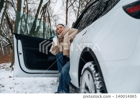 Woman in winter attire is stepping out of a car in a snowy forest, holding a warm drink, showcasing a cozy outdoor experience with nature Woman in winter attire is stepping out of a car in a snowy forest, holding a warm drink, showcasing a cozy outdoor experience with nature 140014865