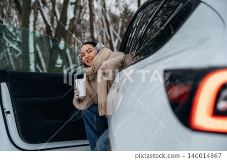 Smiling woman in winter attire holding coffee cup while sitting in car doorway, surrounded by snowy trees, capturing cozy winter vibes and outdoor adventure Smiling woman in winter attire holding coffee cup while sitting in car doorway, surrounded by snowy trees, capturing cozy winter vibes and outdoor adventure 140014867