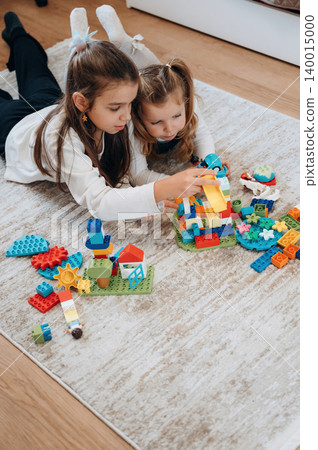 Two young girls are playing together on a soft rug, building colorful structures with toy blocks, showcasing creativity and teamwork in a cozy indoor environment Two young girls are playing together on a soft rug, building colorful structures with toy blocks, showcasing creativity and teamwork in a cozy indoor environment 140015000