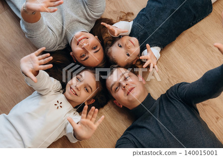 Family enjoying quality time together, lying on wooden floor, smiling and waving hands, capturing joyful moments of togetherness and love Family enjoying quality time together, lying on wooden floor, smiling and waving hands, capturing joyful moments of togetherness and love 140015001