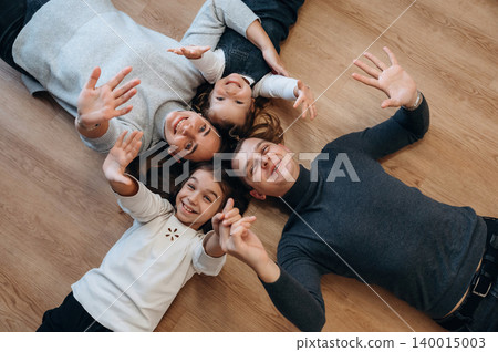 Family enjoying quality time together, lying on wooden floor, smiling and waving hands, capturing joyful moments of togetherness and happiness Family enjoying quality time together, lying on wooden floor, smiling and waving hands, capturing joyful moments of togetherness and happiness 140015003