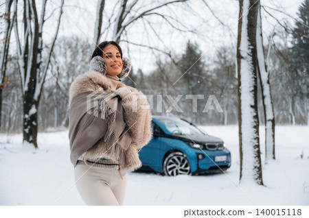 Young woman with dark hair, wearing a stylish fur coat, smiles joyfully in a snowy landscape, with a blue car parked nearby, capturing the essence of winter fashion and outdoor adventure Young woman with dark hair, wearing a stylish fur coat, smiles joyfully in a snowy landscape, with a blue car parked nearby, capturing the essence of winter fashion and outdoor adventure 140015118