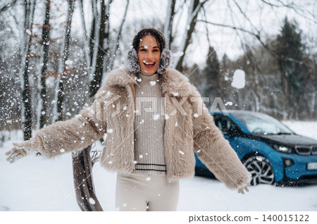 Smiling woman in a fluffy winter coat enjoys snowfall outdoors, with a blue electric car parked nearby, capturing the joy of winter and the beauty of nature Smiling woman in a fluffy winter coat enjoys snowfall outdoors, with a blue electric car parked nearby, capturing the joy of winter and the beauty of nature 140015122