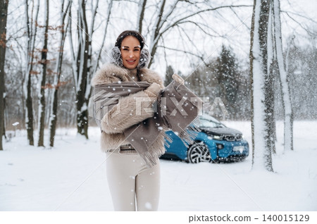 Elegant woman in winter attire, smiling while holding a warm scarf, surrounded by falling snowflakes in a serene snowy landscape with a blue vehicle in the background Elegant woman in winter attire, smiling while holding a warm scarf, surrounded by falling snowflakes in a serene snowy landscape with a blue vehicle in the background 140015129