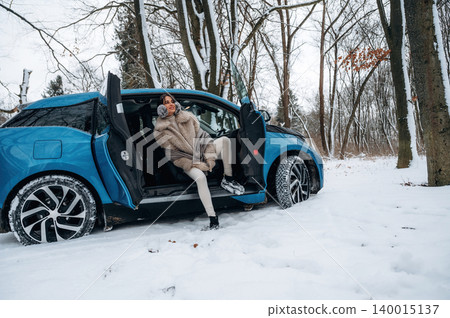 Woman in a stylish fur coat is sitting on the edge of a blue car parked in a snowy forest, showcasing winter fashion and the beauty of nature in a serene environment Woman in a stylish fur coat is sitting on the edge of a blue car parked in a snowy forest, showcasing winter fashion and the beauty of nature in a serene environment 140015137