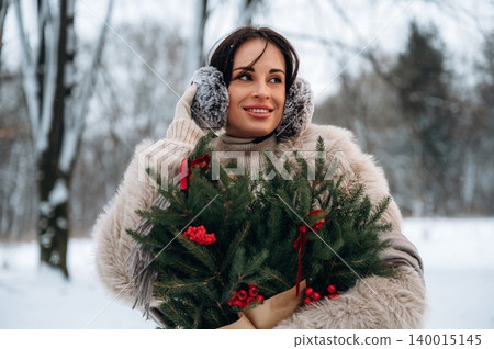 Woman in winter attire, holding a festive evergreen arrangement with red berries, smiles joyfully in a snowy outdoor setting, embodying holiday spirit and warmth 140015145