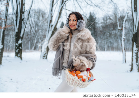 Woman in cozy winter attire, holding a basket of oranges, walking through a snowy landscape, surrounded by trees, showcasing the beauty of winter and seasonal harvest Woman in cozy winter attire, holding a basket of oranges, walking through a snowy landscape, surrounded by trees, showcasing the beauty of winter and seasonal harvest 140015174