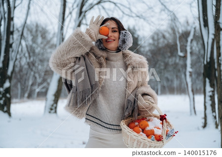 Woman in winter attire, holding a basket of oranges, smiles joyfully in a snowy landscape, surrounded by trees, embodying the spirit of winter festivities and seasonal cheer 140015176
