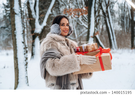 Woman with dark hair wearing winter attire holds beautifully wrapped gifts in snowy forest, showcasing festive spirit and holiday joy in a serene winter landscape 140015195