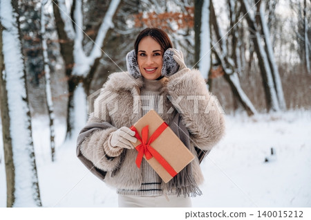 Woman with dark hair wearing a cozy fur coat and earmuffs, holding a gift box with a red ribbon, standing in a snowy forest, celebrating winter holidays with joy and warmth 140015212