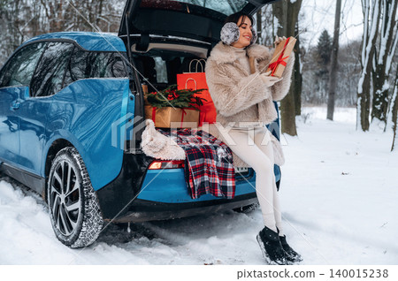 Woman in winter attire sits on the back of a blue car, holding a gift, surrounded by festive decorations and snow-covered trees, celebrating the holiday spirit Woman in winter attire sits on the back of a blue car, holding a gift, surrounded by festive decorations and snow-covered trees, celebrating the holiday spirit 140015238