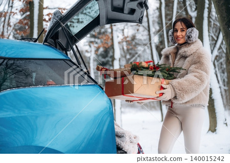 Woman in winter attire, carrying festive gifts from a blue car trunk in a snowy forest, showcasing holiday spirit and seasonal joy with nature backdrop Woman in winter attire, carrying festive gifts from a blue car trunk in a snowy forest, showcasing holiday spirit and seasonal joy with nature backdrop 140015242