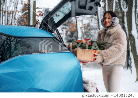 Woman in winter attire, wearing earmuffs, is unloading a gift box from a blue car trunk in a snowy forest, showcasing holiday spirit and seasonal joy Woman in winter attire, wearing earmuffs, is unloading a gift box from a blue car trunk in a snowy forest, showcasing holiday spirit and seasonal joy 140015243
