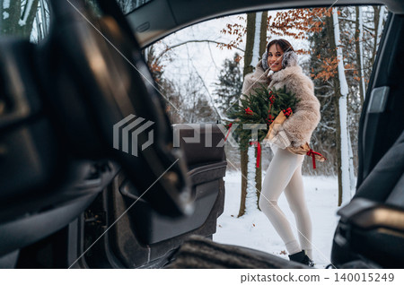 Woman in winter attire holding a festive evergreen arrangement stands beside a car in a snowy forest, capturing the essence of holiday spirit and seasonal joy Woman in winter attire holding a festive evergreen arrangement stands beside a car in a snowy forest, capturing the essence of holiday spirit and seasonal joy 140015249
