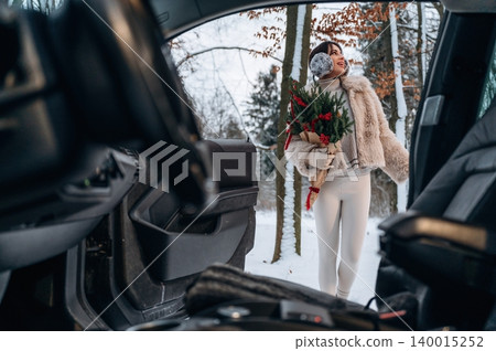 Woman in winter attire holding festive bouquet stands outside car in snowy landscape, showcasing holiday spirit and joy in a serene outdoor environment with nature Woman in winter attire holding festive bouquet stands outside car in snowy landscape, showcasing holiday spirit and joy in a serene outdoor environment with nature 140015252