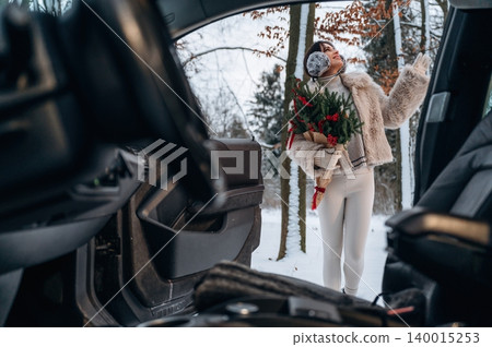 Woman in winter attire, holding festive flowers, stands outside a car in a snowy forest, showcasing a joyful holiday spirit and connection with nature Woman in winter attire, holding festive flowers, stands outside a car in a snowy forest, showcasing a joyful holiday spirit and connection with nature 140015253