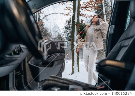 Woman in winter attire, holding bouquet of flowers, steps out of car into snowy landscape, surrounded by trees, embodying festive spirit and seasonal joy Woman in winter attire, holding bouquet of flowers, steps out of car into snowy landscape, surrounded by trees, embodying festive spirit and seasonal joy 140015254
