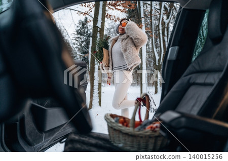 Woman in cozy winter attire stands beside a car in a snowy forest, holding a festive bouquet and a basket, capturing the essence of holiday cheer and nature's beauty Woman in cozy winter attire stands beside a car in a snowy forest, holding a festive bouquet and a basket, capturing the essence of holiday cheer and nature's beauty 140015256