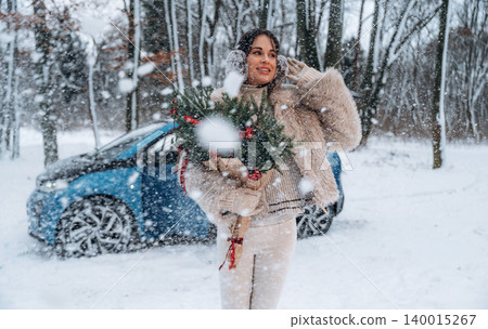 Woman with dark hair wearing a cozy sweater and fur coat holds a festive wreath in snowy landscape, with a blue car parked nearby, celebrating winter holiday spirit Woman with dark hair wearing a cozy sweater and fur coat holds a festive wreath in snowy landscape, with a blue car parked nearby, celebrating winter holiday spirit 140015267