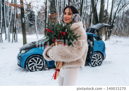 Woman in fluffy winter coat holding festive greenery with red berries, standing near electric car in snowy forest, embodying holiday spirit and seasonal joy Woman in fluffy winter coat holding festive greenery with red berries, standing near electric car in snowy forest, embodying holiday spirit and seasonal joy 140015270