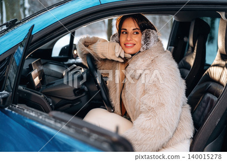 Woman with long dark hair, wearing a cozy fur coat and gloves, is smiling while sitting in a blue car, enjoying a winter day, showcasing warmth and comfort in a cold environment Woman with long dark hair, wearing a cozy fur coat and gloves, is smiling while sitting in a blue car, enjoying a winter day, showcasing warmth and comfort in a cold environment 140015278