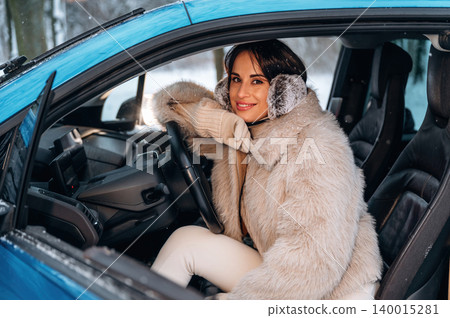 Smiling woman with dark hair wearing a fluffy winter coat and earmuffs, sitting in a blue car, enjoying a snowy day, radiating warmth and joy in a cozy atmosphere Smiling woman with dark hair wearing a fluffy winter coat and earmuffs, sitting in a blue car, enjoying a snowy day, radiating warmth and joy in a cozy atmosphere 140015281