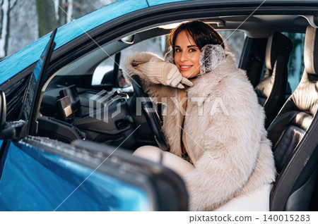 Smiling woman in a cozy fur coat sits in a blue car, showcasing a warm winter atmosphere, with snow-covered trees visible outside the vehicle Smiling woman in a cozy fur coat sits in a blue car, showcasing a warm winter atmosphere, with snow-covered trees visible outside the vehicle 140015283