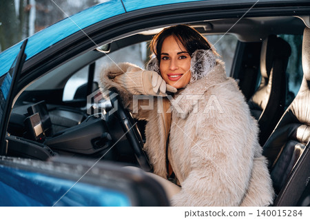 Young woman with long dark hair, wearing a cozy fur coat, is smiling while sitting in a car, showcasing a warm and inviting atmosphere in a winter setting Young woman with long dark hair, wearing a cozy fur coat, is smiling while sitting in a car, showcasing a warm and inviting atmosphere in a winter setting 140015284