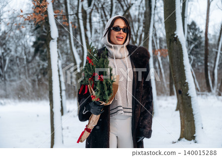 Stylish woman in winter attire, holding a festive bouquet, smiles joyfully in a snowy forest, surrounded by tall trees and a serene winter landscape, celebrating the holiday season Stylish woman in winter attire, holding a festive bouquet, smiles joyfully in a snowy forest, surrounded by tall trees and a serene winter landscape, celebrating the holiday season 140015288