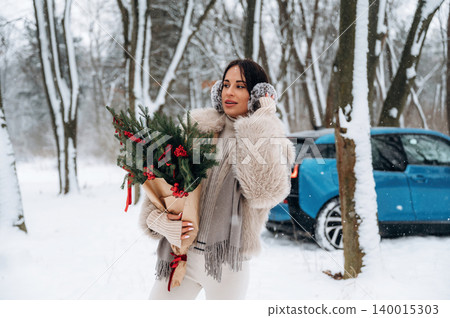 Woman with dark hair, wearing cozy winter attire, holding festive greenery bouquet in snowy forest, with blue car parked nearby, celebrating holiday spirit and winter joy Woman with dark hair, wearing cozy winter attire, holding festive greenery bouquet in snowy forest, with blue car parked nearby, celebrating holiday spirit and winter joy 140015303