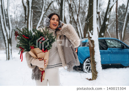 Woman in winter attire, holding festive greenery with red accents, smiles joyfully in a snowy forest setting, with a blue vehicle parked nearby, celebrating the holiday season Woman in winter attire, holding festive greenery with red accents, smiles joyfully in a snowy forest setting, with a blue vehicle parked nearby, celebrating the holiday season 140015304