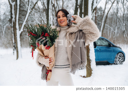 Woman in winter attire, holding a festive bouquet of red berries and greenery, smiles joyfully in a snowy landscape, with a blue vehicle in the background, celebrating the holiday season Woman in winter attire, holding a festive bouquet of red berries and greenery, smiles joyfully in a snowy landscape, with a blue vehicle in the background, celebrating the holiday season 140015305