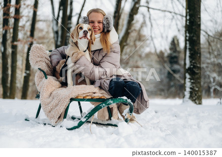 Young woman with long blonde hair wearing winter coat and earmuffs is sitting on a green sled in the snow with a beagle dog during a snowy day in a forested area 140015387