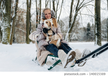 Young girl with blonde hair wearing earmuffs is sitting on a sled in a snowy park holding a beagle dog surrounded by trees during winter leisure activities Young girl with blonde hair wearing earmuffs is sitting on a sled in a snowy park holding a beagle dog surrounded by trees during winter leisure activities 140015389