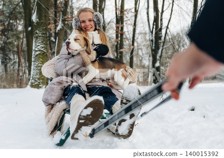 Young girl with long blonde hair wearing earmuffs is sitting on a sled in the snow, holding a beagle dog while a hand is pulling the sled in a winter forest setting 140015392