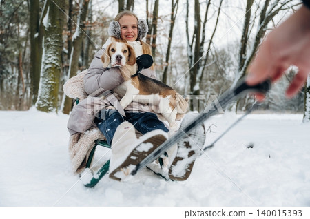 Young woman with blonde hair wearing winter clothing is sitting on a sled in the snow holding a beagle dog while a hand is pulling the sled in a snowy forest Young woman with blonde hair wearing winter clothing is sitting on a sled in the snow holding a beagle dog while a hand is pulling the sled in a snowy forest 140015393