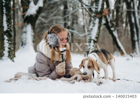 Young woman with blonde hair wearing earmuffs is playing with a beagle dog on a blanket in a snowy forest during winter season with trees in the background Young woman with blonde hair wearing earmuffs is playing with a beagle dog on a blanket in a snowy forest during winter season with trees in the background 140015403
