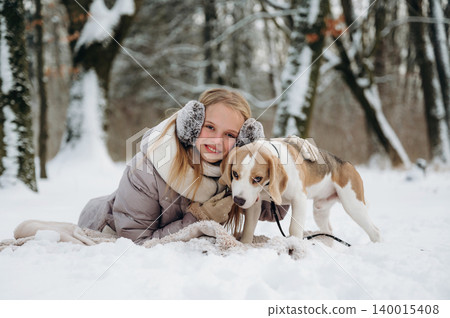 Young girl with blonde hair wearing earmuffs is lying in the snow beside a beagle dog, surrounded by snow-covered trees in a winter forest setting with a cozy blanket 140015408