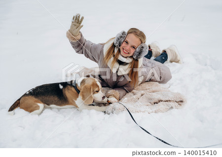 Young girl with long blonde hair wearing earmuffs and gloves is lying in the snow with a beagle dog, enjoying a winter day outdoors on a blanket in a snowy landscape Young girl with long blonde hair wearing earmuffs and gloves is lying in the snow with a beagle dog, enjoying a winter day outdoors on a blanket in a snowy landscape 140015410
